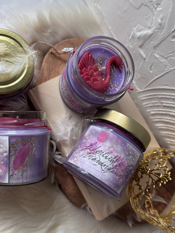 Das Bild zeigt die Buchkerze "Federlicht und Tränenglanz". Auf dem rosa und goldenen Label steht mittig in geschwungenen Buchstaben der Titel. Unter dem goldenen Schraubdeckel hängt an einem pinken Band ein silberner Schwanenanhänger. Der Wachs ist lila-weiß mit goldenem Glitter, mit einer pinken Feder neben dem Label. Um den Docht herum ist ein pinker Schwan aus Wachs.